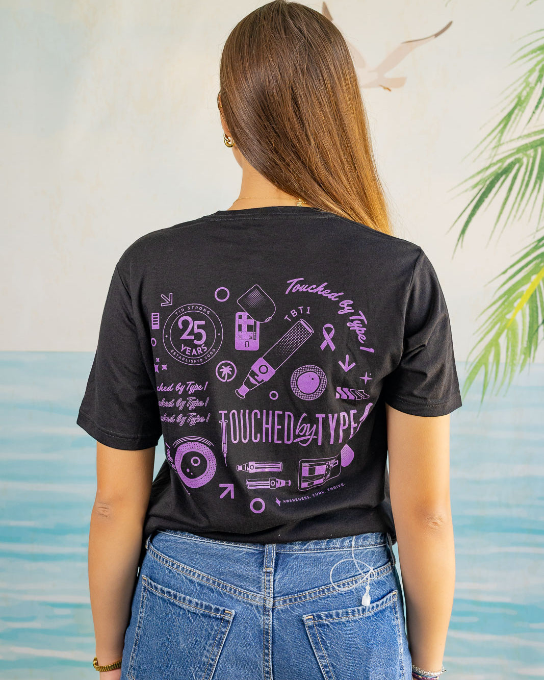 Person wearing a black t-shirt with purple graphics and text, standing against a light-colored wall with greenery.