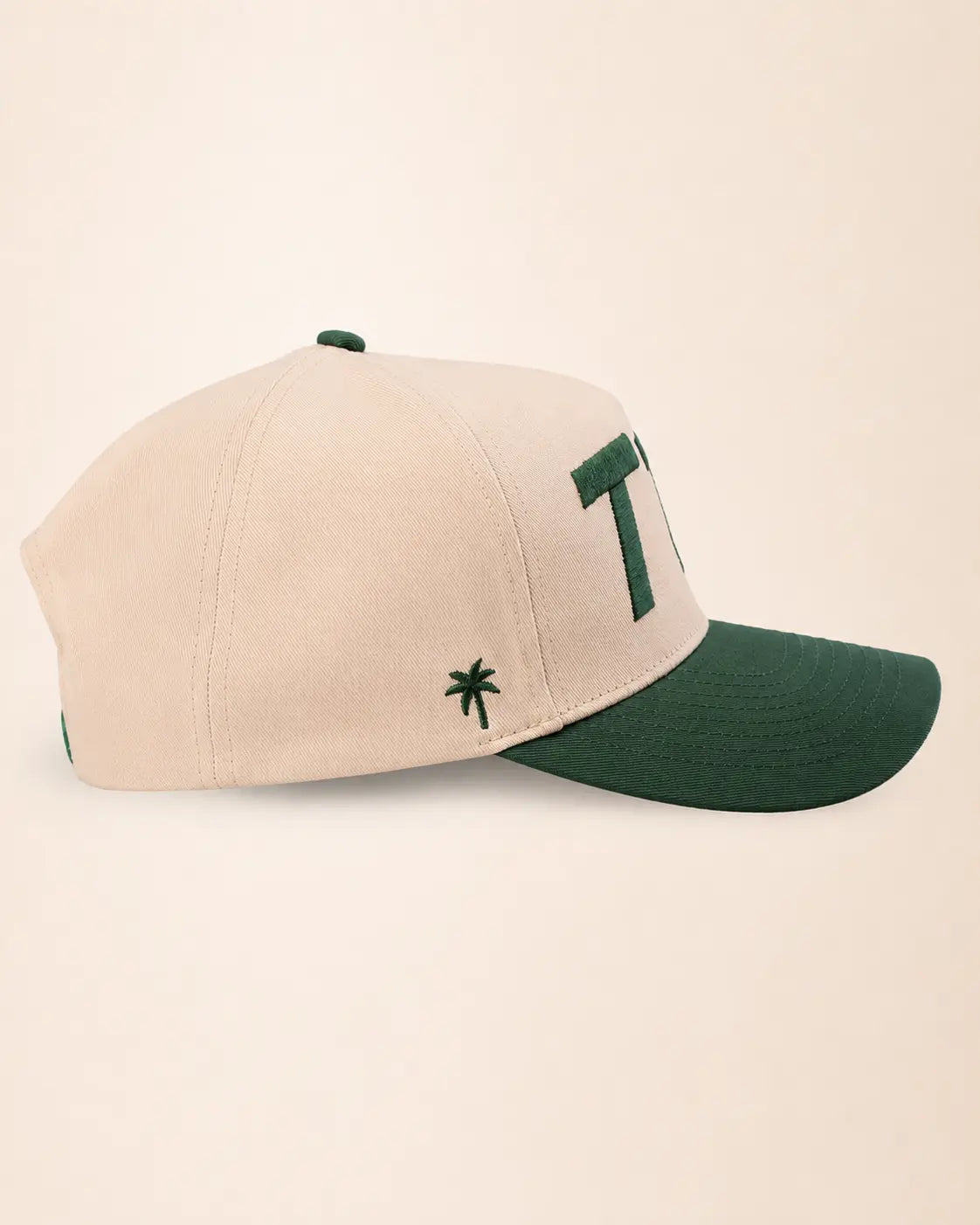 A structured baseball cap in cream and dark green, featuring a large green embroidered letter 'T1d' on the side and a small green palm tree logo on the front, set against a plain cream background.