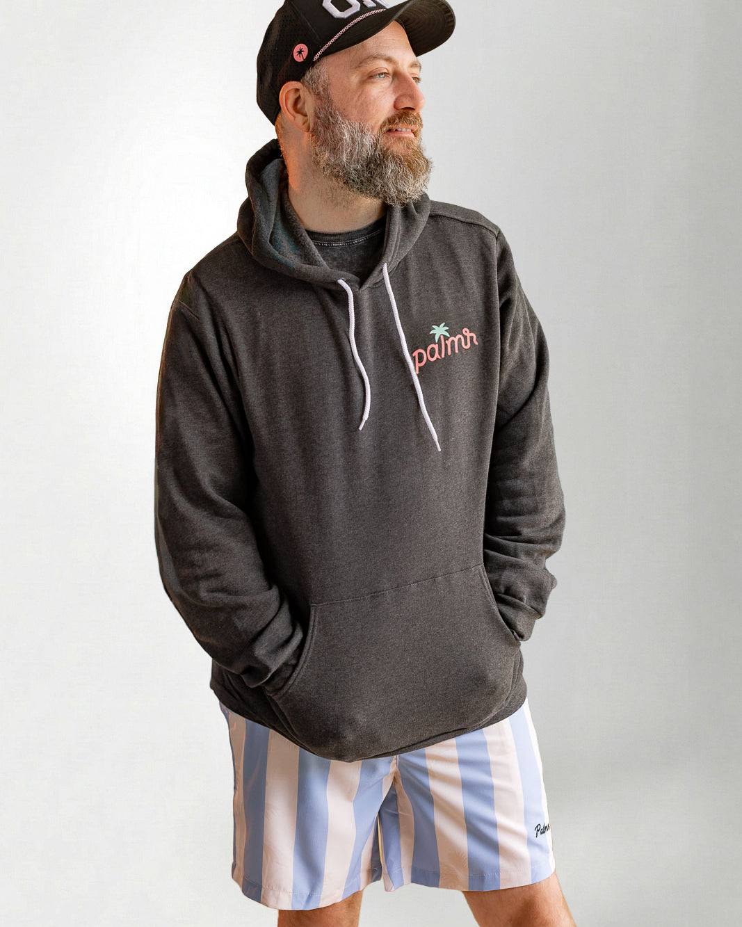Man wearing a dark hoodie with a logo, striped shorts, and a cap on a plain background