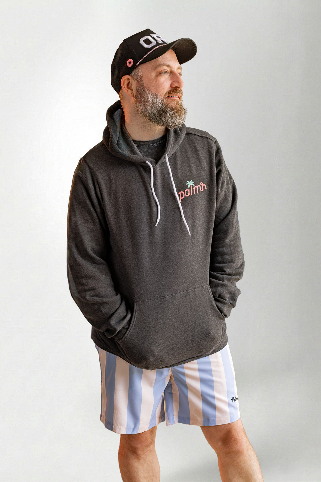 Man wearing a dark hoodie with a logo, striped shorts, and a cap on a plain background