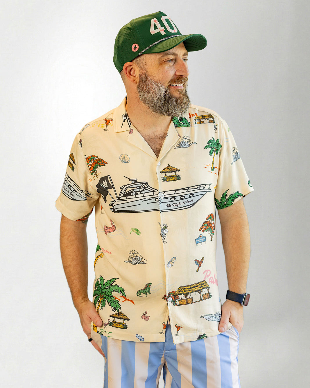 Man wearing a patterned shirt and striped shorts with a cap on a plain background