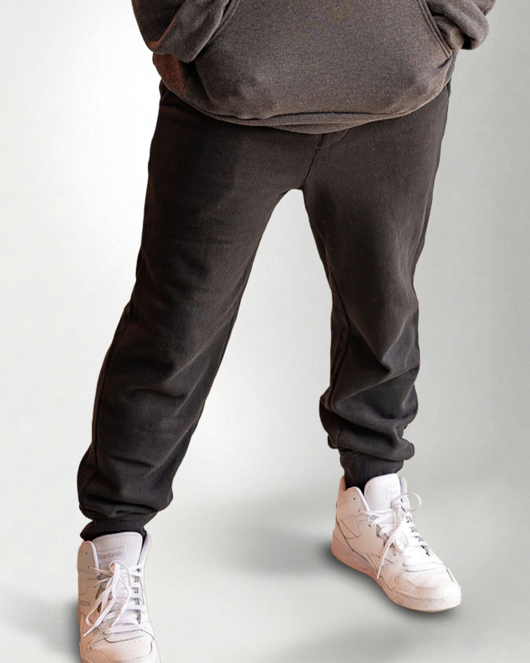 Person wearing a dark gray hoodie and pants with white sneakers on a light gray background