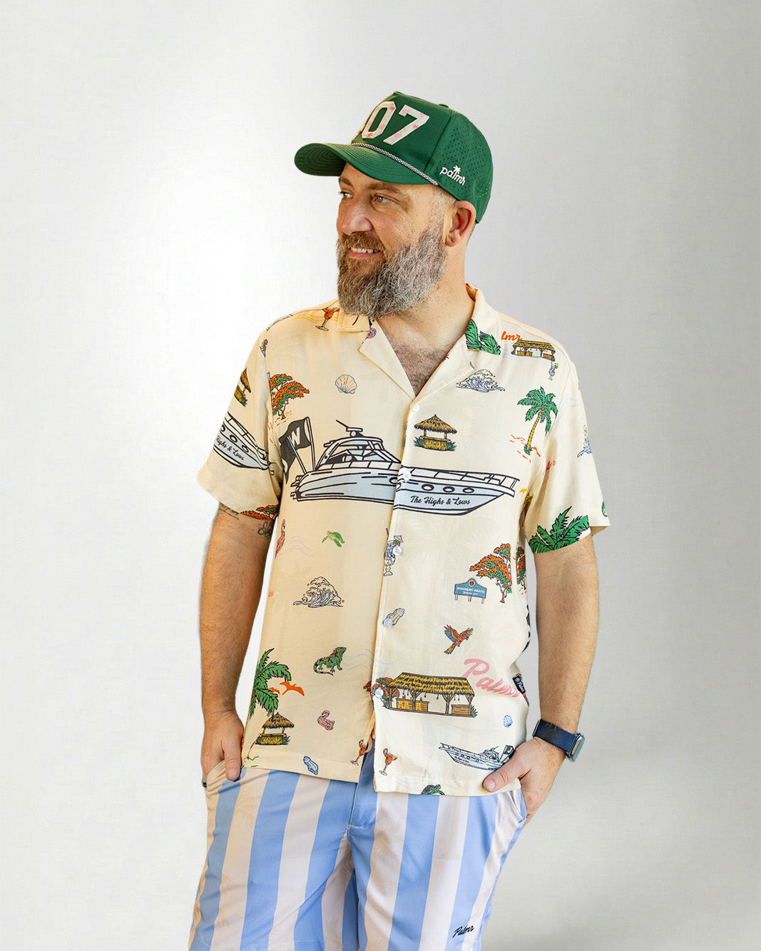 Man wearing a patterned shirt and striped shorts with a cap on a plain background