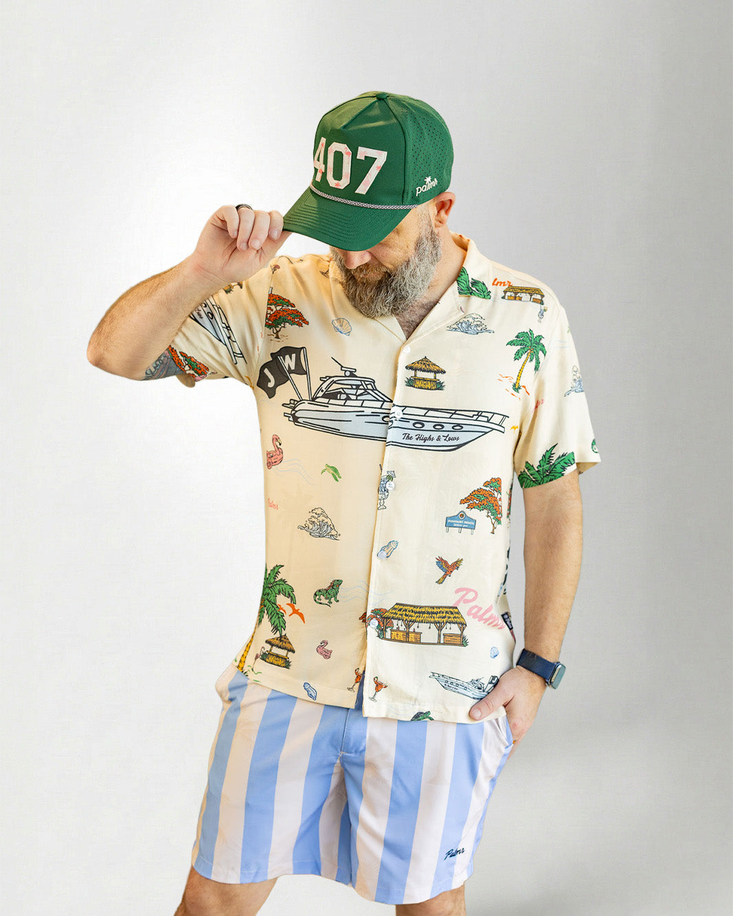 Man wearing a patterned shirt and striped shorts with a cap on a plain background
