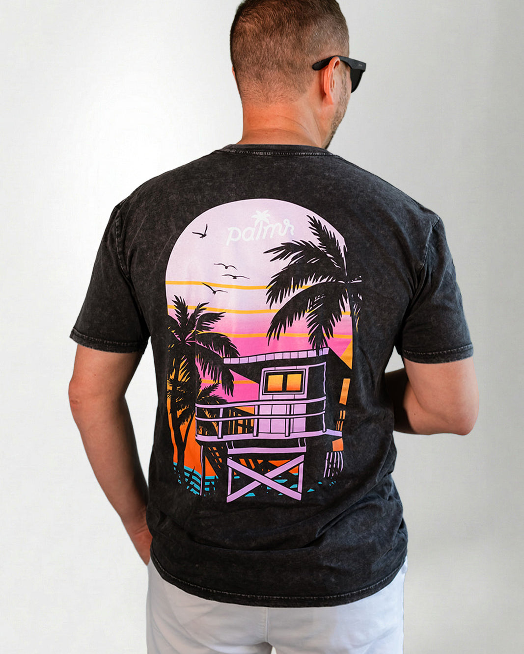 Man wearing a black t-shirt with a colorful beach house design on the back, standing against a plain background.