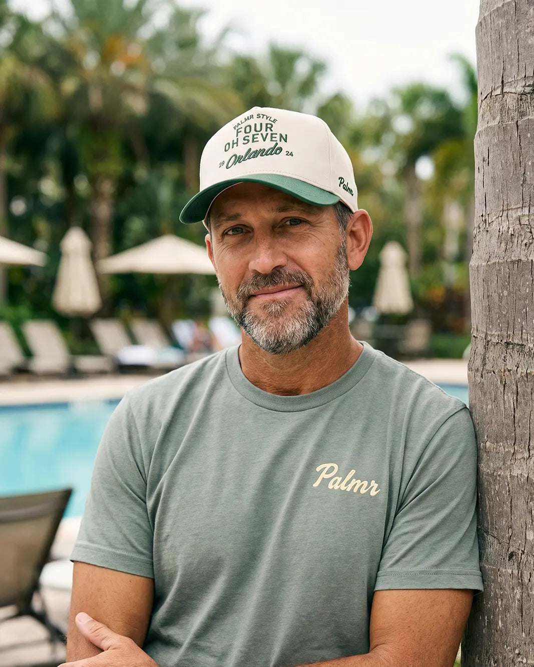 A man leaning against a palm tree poolside, wearing a light green t-shirt with 'Palmr' embroidered on the chest and a beige baseball cap with green text that reads 'FOUR OR SEVEN Orlando'.