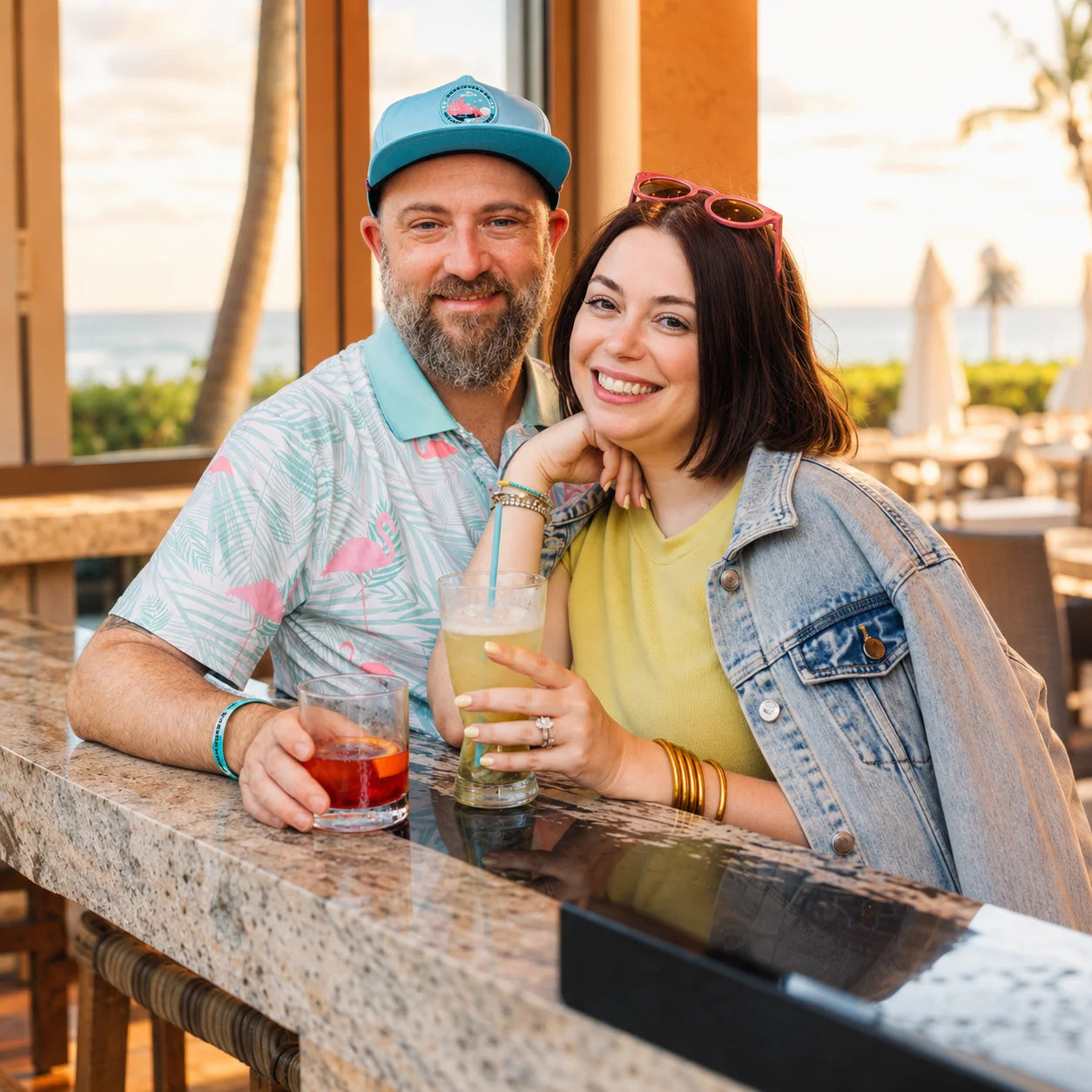 Man and woman sitting at a bar counter with drinks, smiling at the camera.