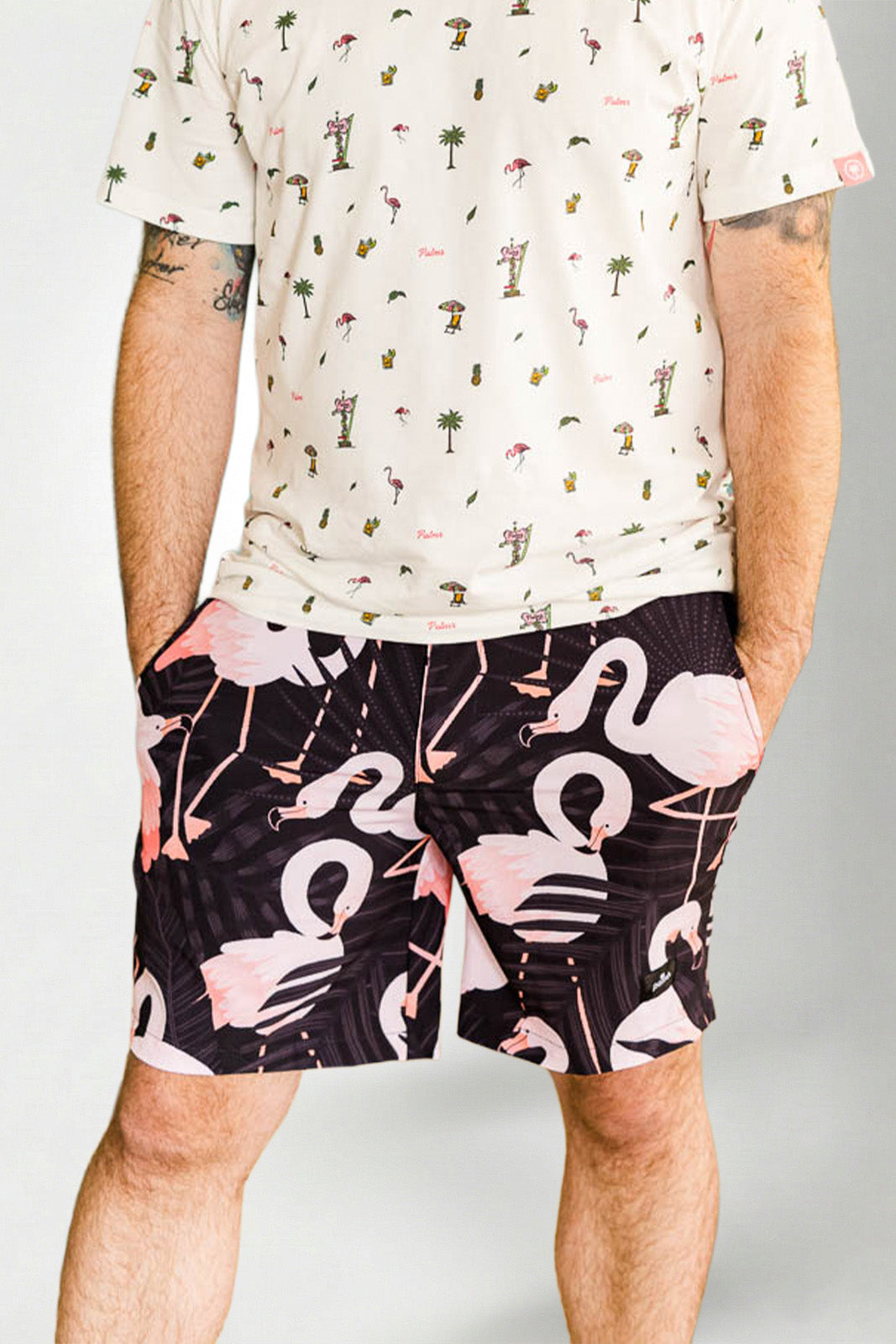 Person wearing a white shirt with a palm tree pattern and black shorts with pink flamingo pattern on a plain background