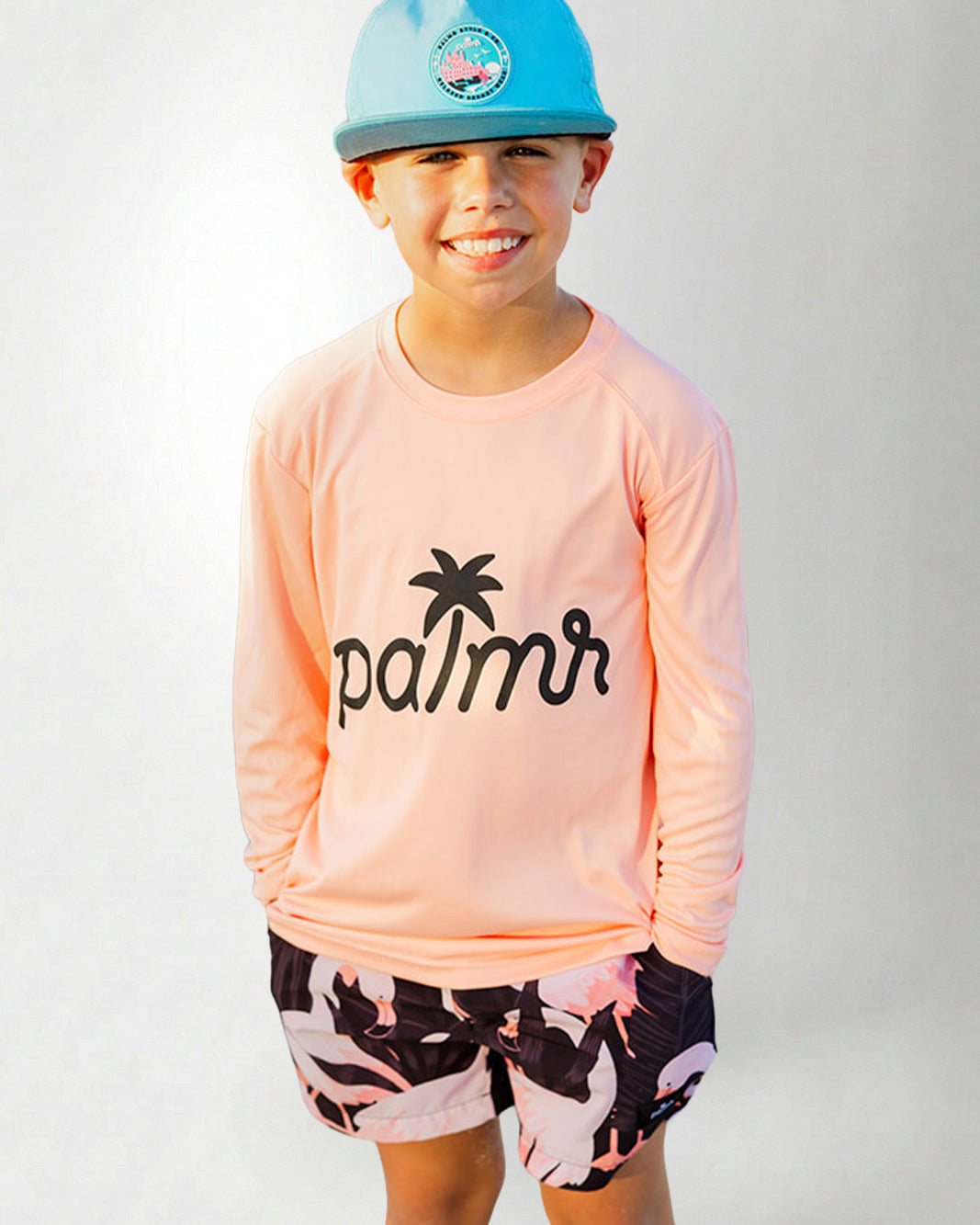 Child wearing a pink long-sleeve shirt with 'palmr' logo and camouflage shorts, smiling against a plain background.