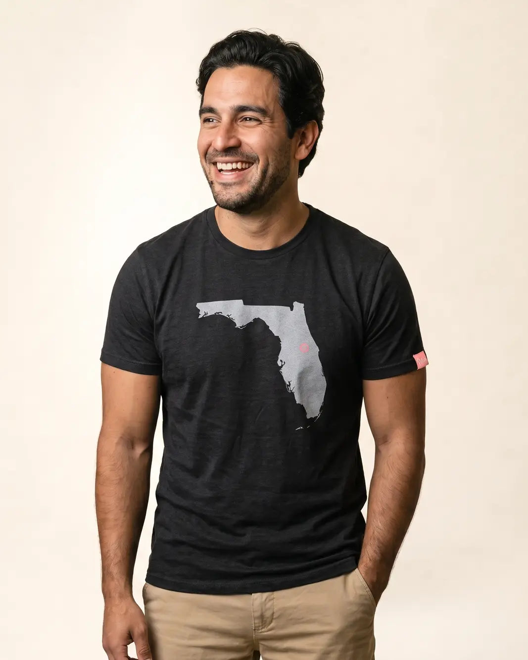A man smiling and looking to the side, wearing a black crewneck t-shirt with a gray silhouette graphic of the state of Florida on the front, paired with khaki pants, against a plain beige background.