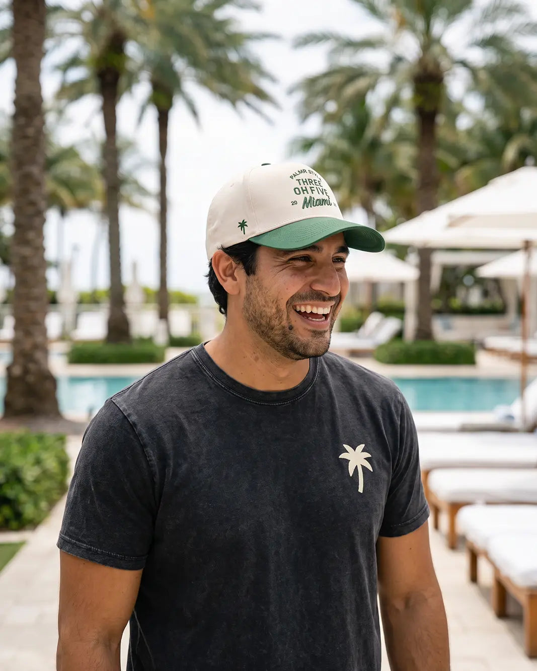 A man smiling outdoors by a pool wearing a dark charcoal t-shirt with a small white palm tree logo on the chest and a cream and green baseball cap with 'THINGS OFTEN Miami' printed on the front.
