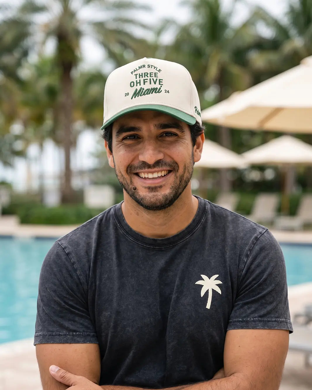 A smiling man wearing a dark charcoal t-shirt with a white palm tree logo and a cream baseball cap with green text, posed outdoors in front of a swimming pool with palm trees and umbrellas in the background.