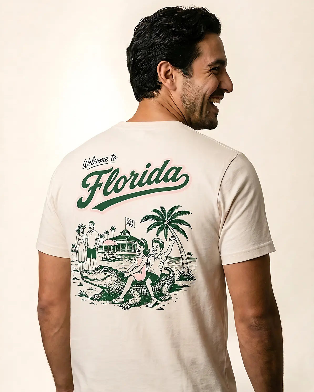 A man shown from the back wearing a cream-colored t-shirt with a green graphic print that reads 'Welcome to Florida' and features a retro illustration of a beach scene with palm trees, a house, and an alligator.