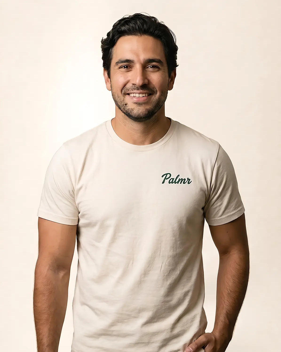 A smiling man wearing a light beige crewneck t-shirt with the brand name 'Palmr' printed in dark green script on the left chest, set against a plain beige background.
