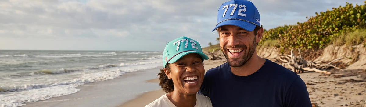 Couple in Palmr 772 Signature hats, teal and blue, on a Treasure Coast beach with sea grapes and dunes behind them