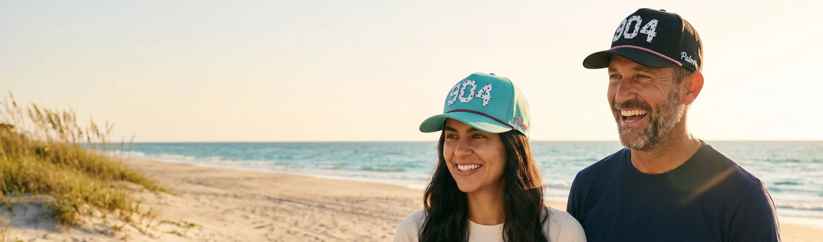 Couple in Palmr 904 Signature hats, teal and black, on a dune-backed Atlantic beach on Jacksonville's First Coast