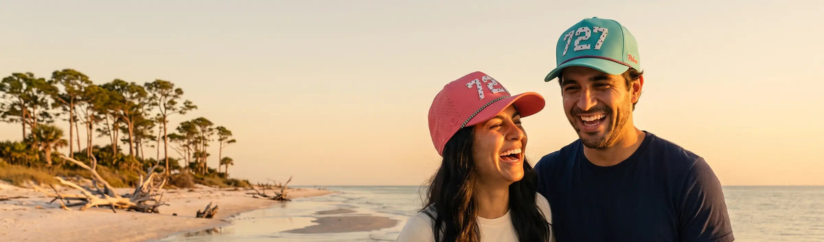 Couple in Palmr 727 Signature hats, pink and teal, on a driftwood beach at sunset on the Pinellas Gulf coast