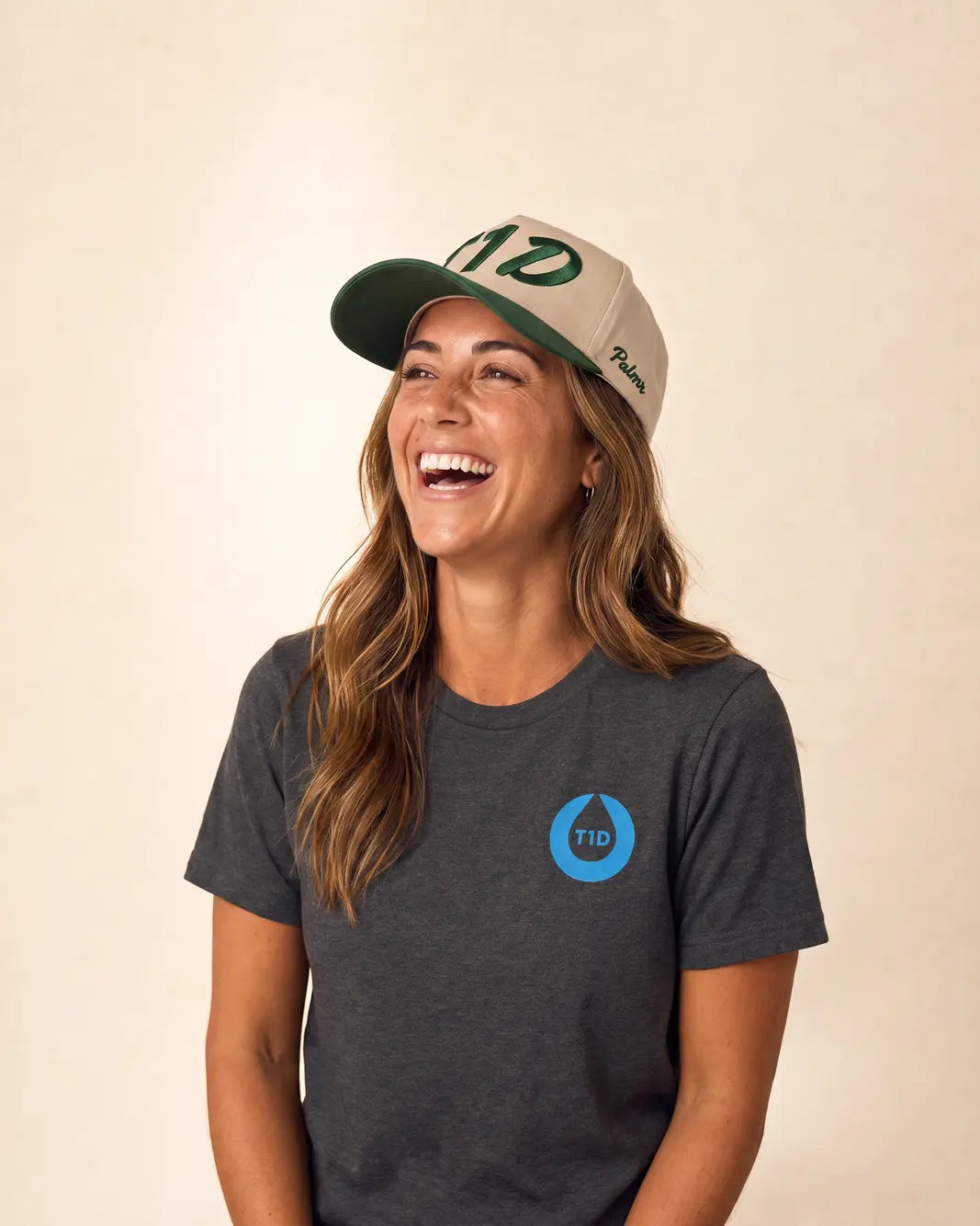 A woman wearing a dark heather gray t-shirt with a blue TID logo on the chest and a beige and green baseball cap, smiling against a plain beige background.