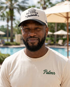 A man wearing a cream-colored t-shirt with 'Palmr' printed in green on the chest and a matching cream trucker hat, standing in front of a blurred resort poolside background.