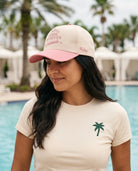 A woman wearing a cream and pink baseball cap with pink embroidered text and a cream t-shirt with a green palm tree logo, standing in front of a resort pool with palm trees in the background.