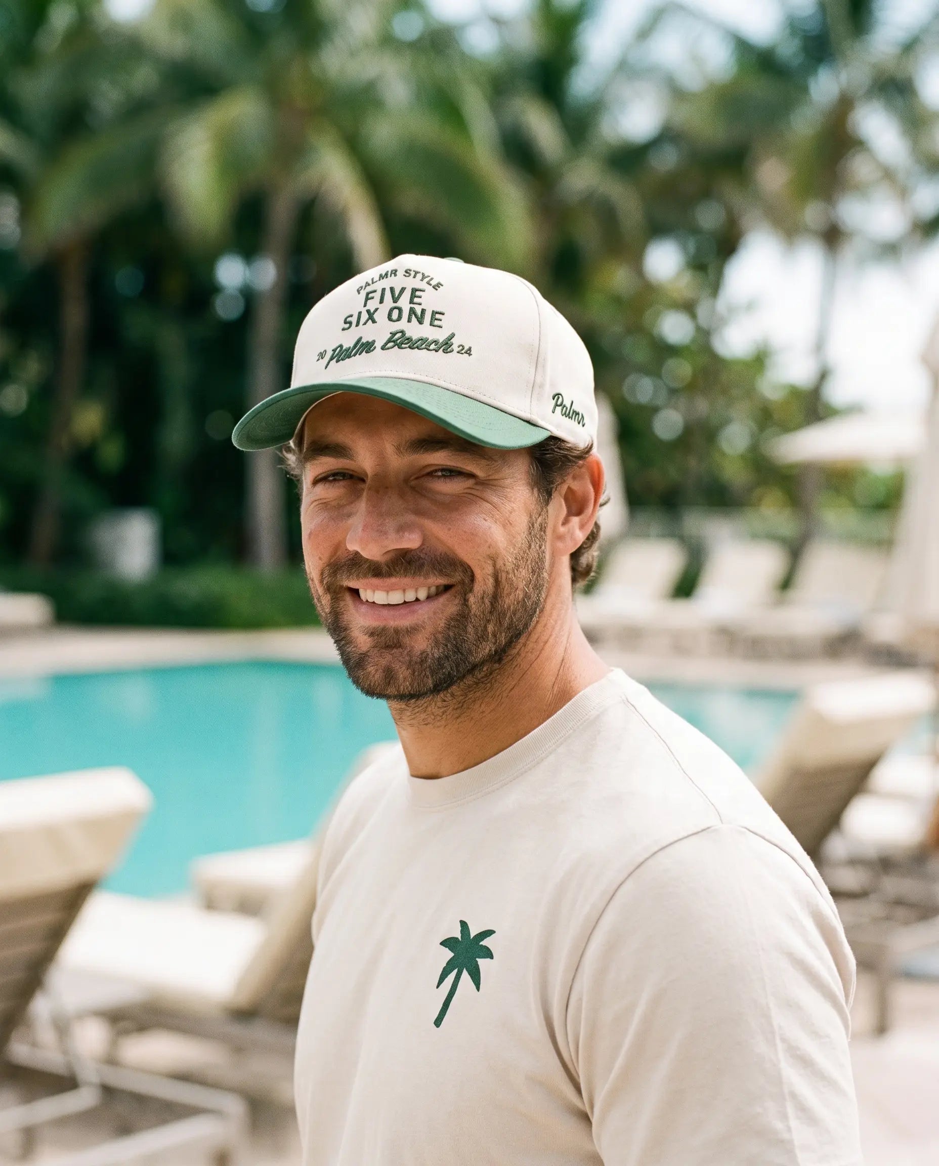 A smiling man wearing a cream-colored baseball cap with green lettering that reads 'FIVE SIX ONE Palm Beach FL' and a matching cream t-shirt with a green palm tree logo, standing in front of a tropical pool with palm trees in the background.