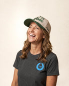 A woman smiling and looking upward, wearing a dark heather gray TID t-shirt with a blue logo on the chest and a beige and green TID 'This Wave' cap. Shot against a solid light beige background.