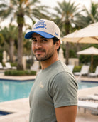 A man wearing a beige and blue trucker hat with 'Four 14 Seven Colorado' printed on it, standing in front of a resort pool with palm trees and lounge chairs.