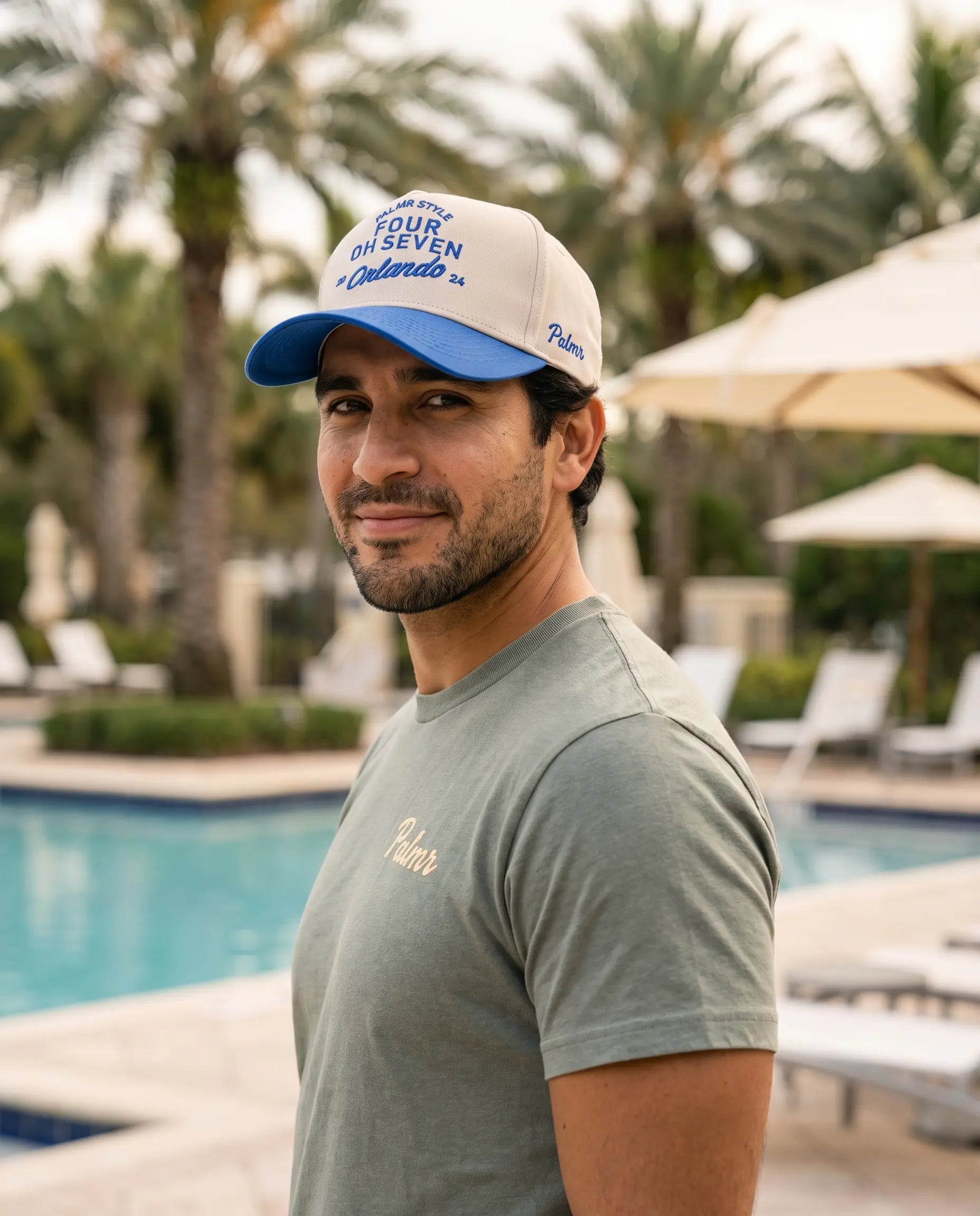 A man wearing a beige and blue trucker hat with 'Four 14 Seven Colorado' printed on it, standing in front of a resort pool with palm trees and lounge chairs.