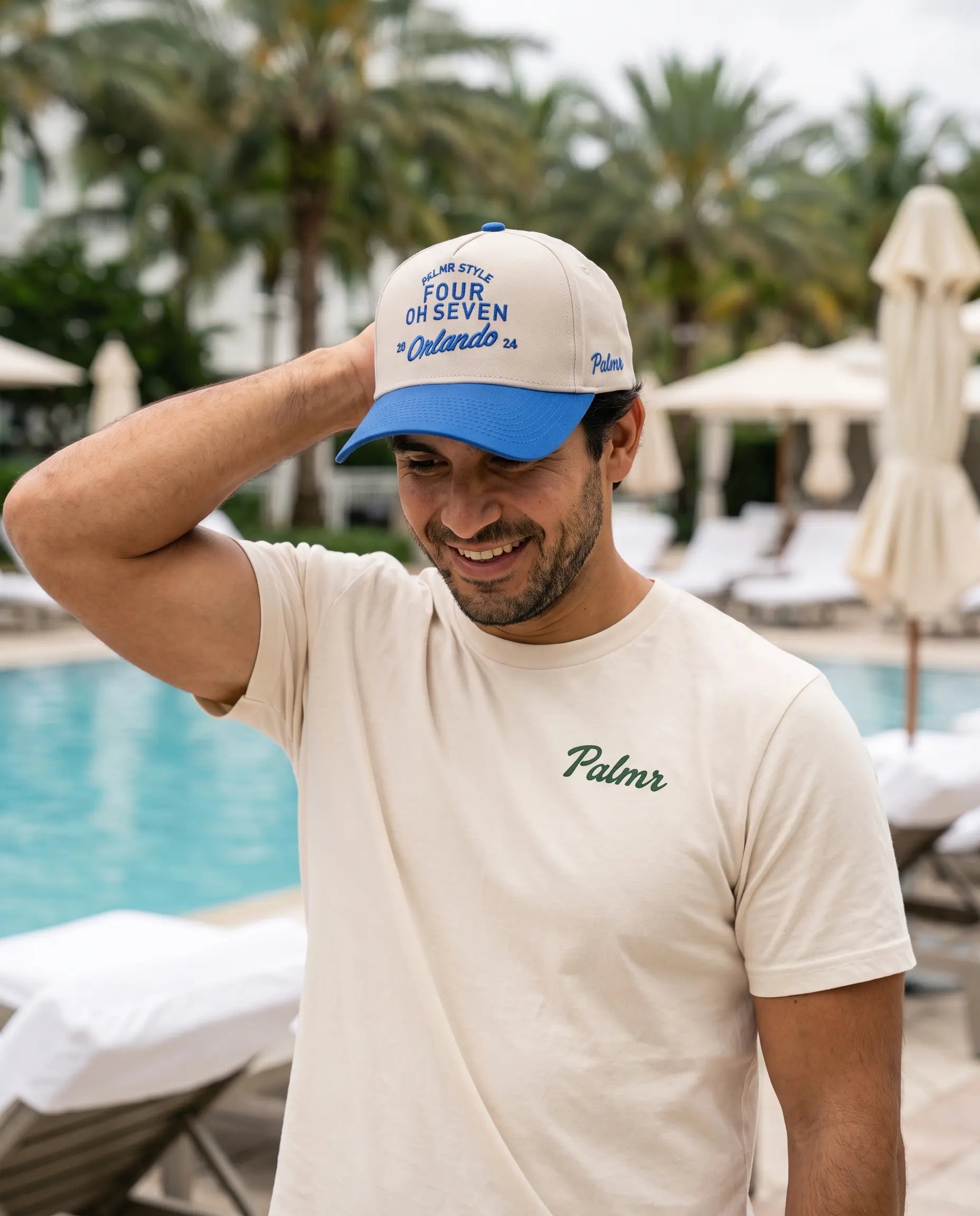 A man wearing a cream t-shirt and a blue and cream baseball cap with 'Palmr' branding, standing by a swimming pool with lounge chairs and palm trees in the background.