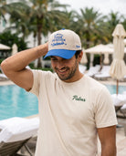 A man wearing a cream t-shirt and a blue and cream baseball cap with 'Palmr' branding, standing by a swimming pool with lounge chairs and palm trees in the background.