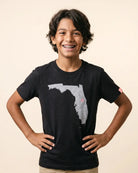 A smiling young boy with dark curly hair and braces wearing a black t-shirt with a gray Florida state graphic on the front, standing against a solid beige background.