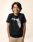 A smiling young boy with dark hair and braces wearing a black t-shirt with a gray Florida state graphic and a small pink dot, paired with khaki pants, against a solid beige background.