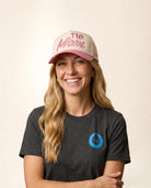 A smiling woman wearing a beige and pink trucker hat with 'TID Mom' written on it, paired with a dark gray TID t-shirt, against a plain beige background.