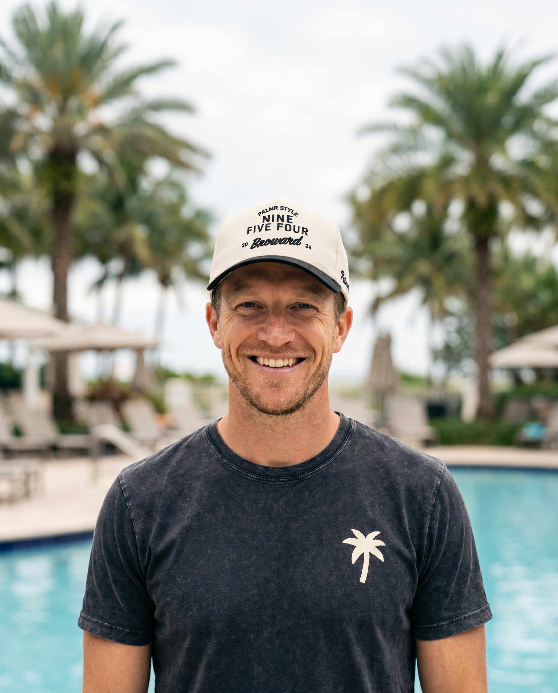 A smiling man standing in front of a swimming pool with palm trees in the background, wearing a white cap with black text and a dark charcoal t-shirt with a small white palm tree logo.
