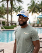 A man wearing a light green t-shirt with a gold embroidered 'Palmer' logo and a cream and blue baseball cap stands in front of a swimming pool with palm trees in the background.