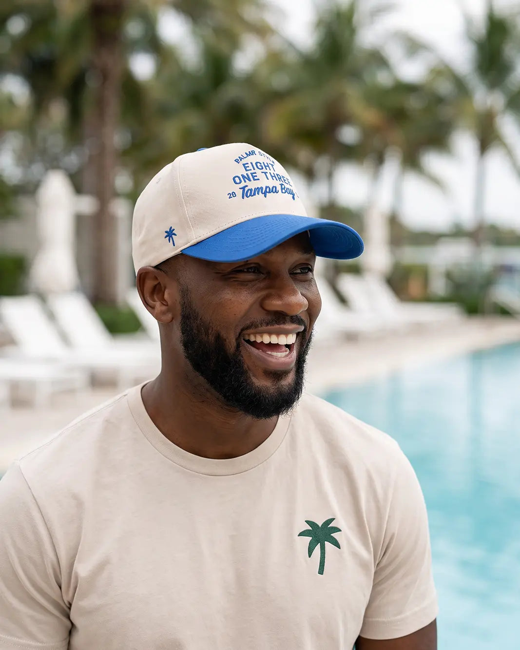 A man smiling outdoors by a swimming pool wearing a beige T-shirt with a green palm tree logo and a beige-and-blue baseball cap with blue text reading 'Welcome to One Tampa Bay'.