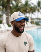 A man smiling outdoors by a swimming pool wearing a beige T-shirt with a green palm tree logo and a beige-and-blue baseball cap with blue text reading 'Welcome to One Tampa Bay'.