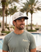 A man wearing the cream and navy 321 Space Coast Heritage Hat with a muted sage green t-shirt featuring 'Palmr' in gold script, set against a blurred background of palm trees and a swimming pool.