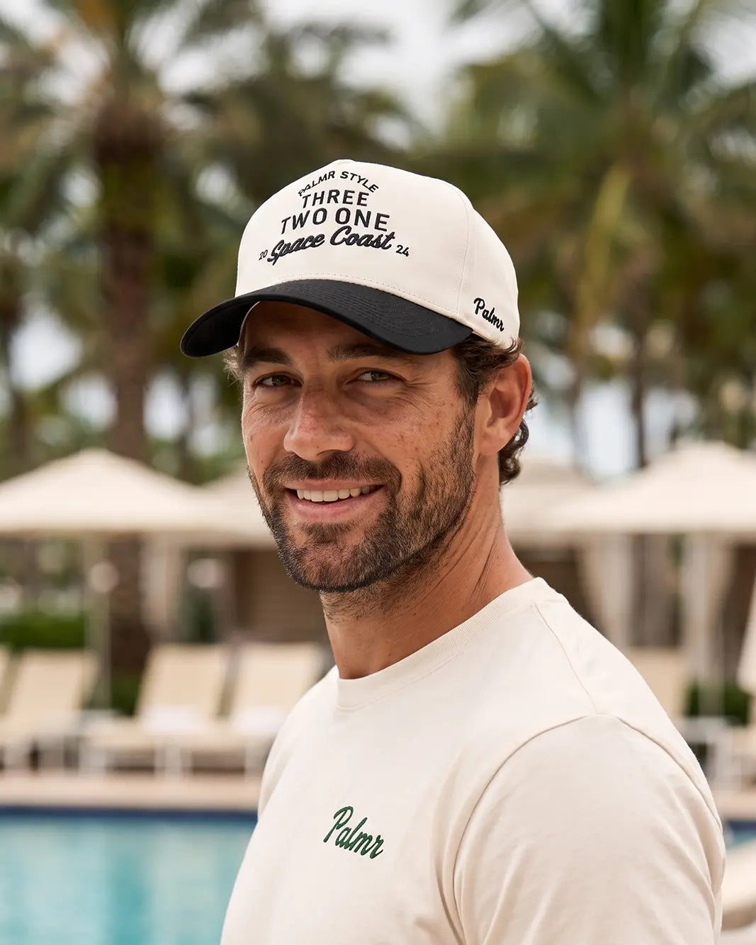 A smiling man wearing a white and navy blue baseball cap with 'Three Two One Square Cocktail' printed on the front, alongside a white t-shirt with 'Palmer' embroidered on the chest, set against a blurred resort background with palm trees and a pool.