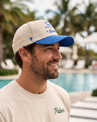 A man wearing a beige and blue baseball cap with 'The Happy Five' and 'Breezeway' branding, standing outdoors near a swimming pool with palm trees in the background.