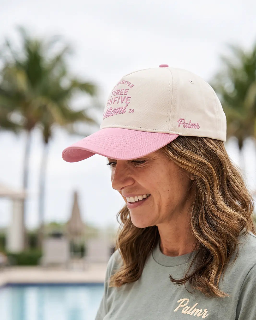 A woman wearing a cream and pink baseball cap with 'Three Five Miami' embroidered on the front and 'Palmr' on the side, paired with a matching light green t-shirt with 'Palmr' embroidered on the chest. The background is a soft-focus outdoor scene with palm trees and a pool.