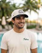 A smiling man wearing a cream-colored t-shirt with 'Palmr' printed in green on the chest and a matching cream cap with black text, set against a blurred background of a pool and palm trees.