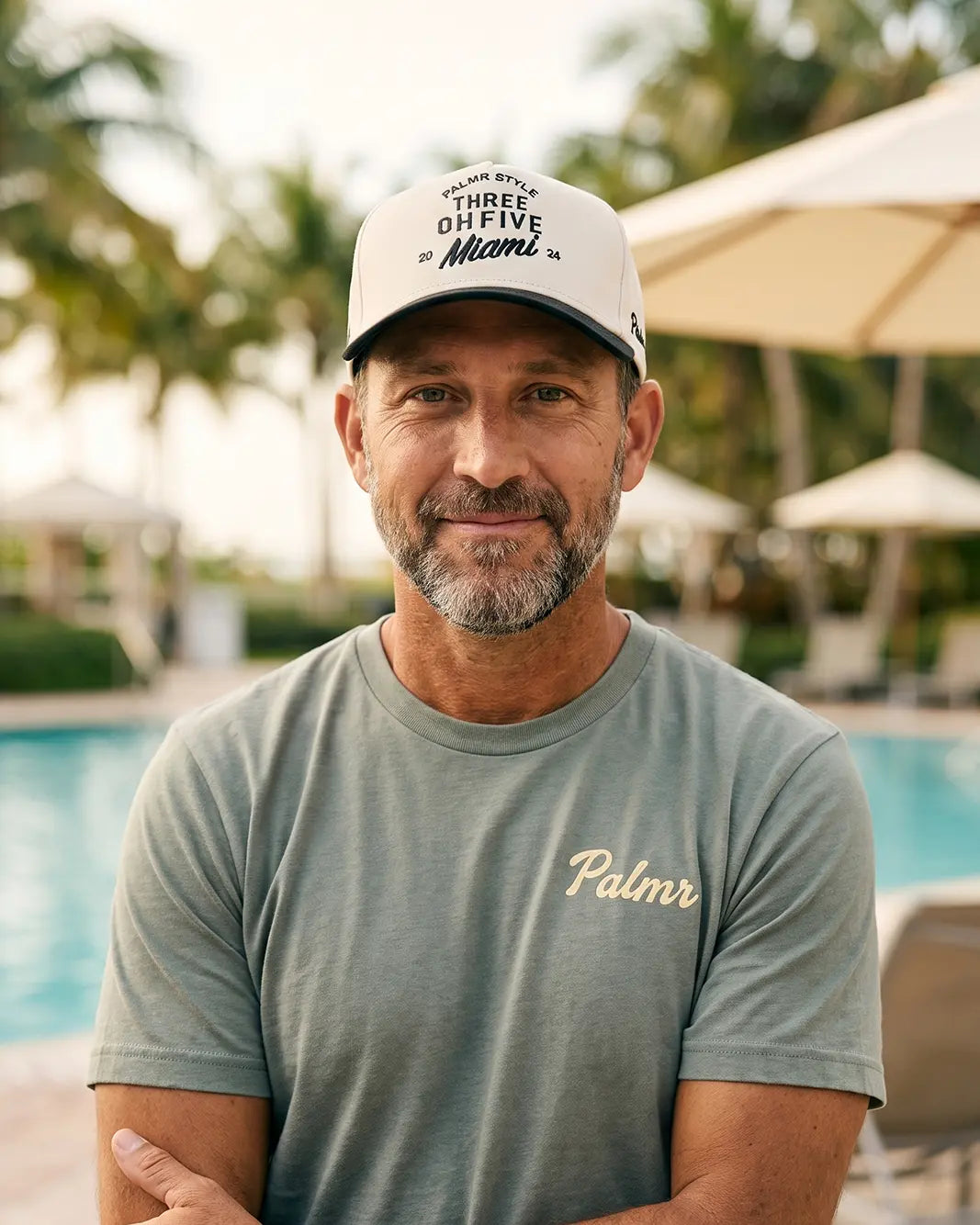 A man wearing a gray t-shirt with 'Palmr' printed on the chest and a white baseball cap with 'THREE GIFTIVE of Miami' text, standing in front of a blurred resort pool background.