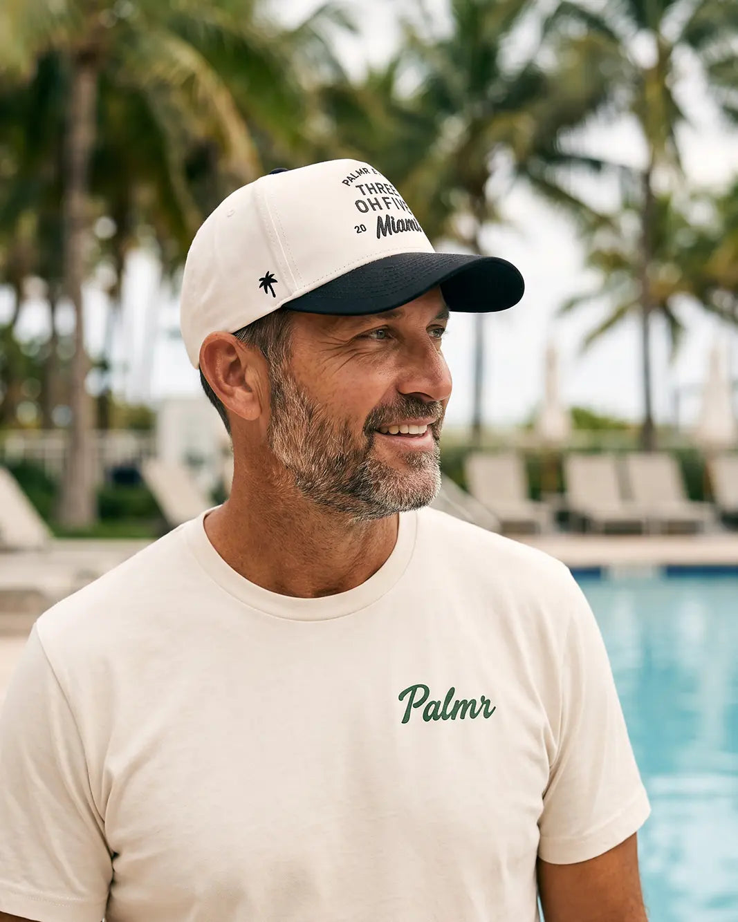 A smiling man wearing a cream t-shirt with 'Palmr' embroidered on the chest and a matching cream and black baseball cap, standing in front of a pool with palm trees in the background.