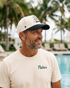 A smiling man wearing a cream t-shirt with 'Palmr' embroidered on the chest and a matching cream and black baseball cap, standing in front of a pool with palm trees in the background.