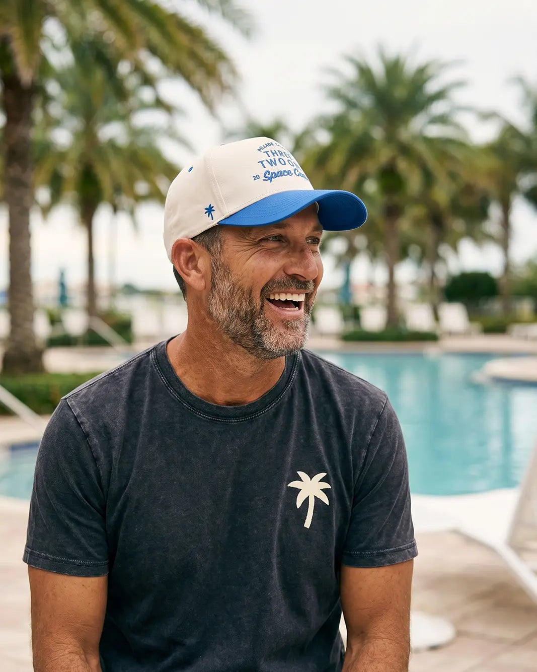 A man wearing a white and blue baseball cap with blue text and a dark gray t-shirt with a white palm tree graphic, sitting in front of a swimming pool with palm trees in the background.