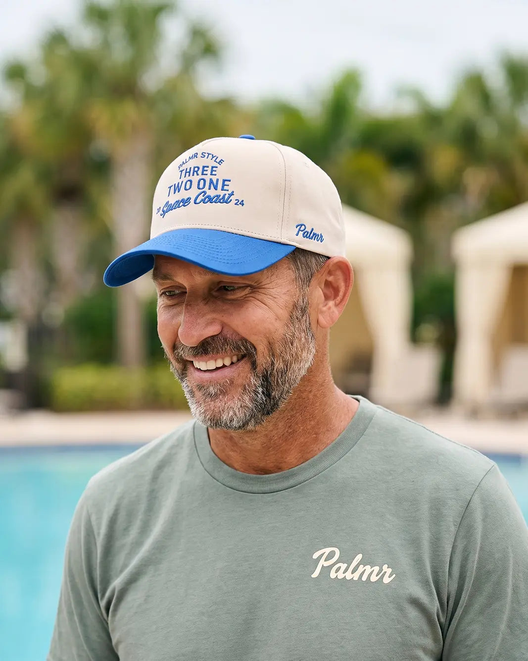 A smiling man wearing a blue and off-white baseball cap with 'Three Two One Squares Coast 50' and 'Palm' branding, paired with a muted green t-shirt that has 'Palm' embroidered on the chest, set against a blurred resort background with a pool and palm trees.