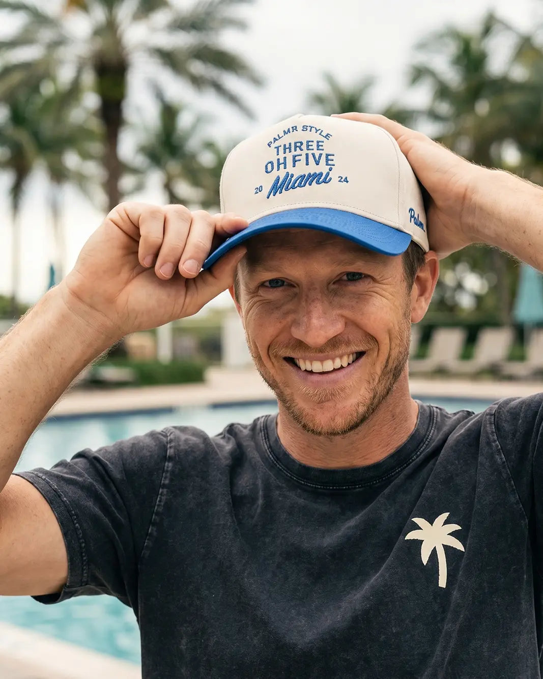 A smiling man wearing a white and blue trucker hat with 'Three Oh Five Miami' text, posing by a swimming pool with palm trees in the background.