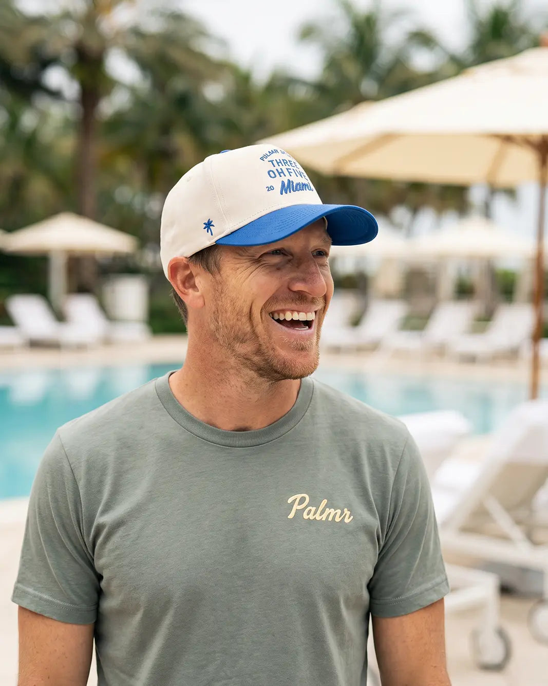 A man smiling outdoors by a swimming pool, wearing a white and blue baseball cap with blue text and a gray t-shirt with the name 'Palmr' embroidered in gold on the chest.