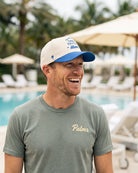 A man smiling outdoors by a swimming pool, wearing a white and blue baseball cap with blue text and a gray t-shirt with the name 'Palmr' embroidered in gold on the chest.